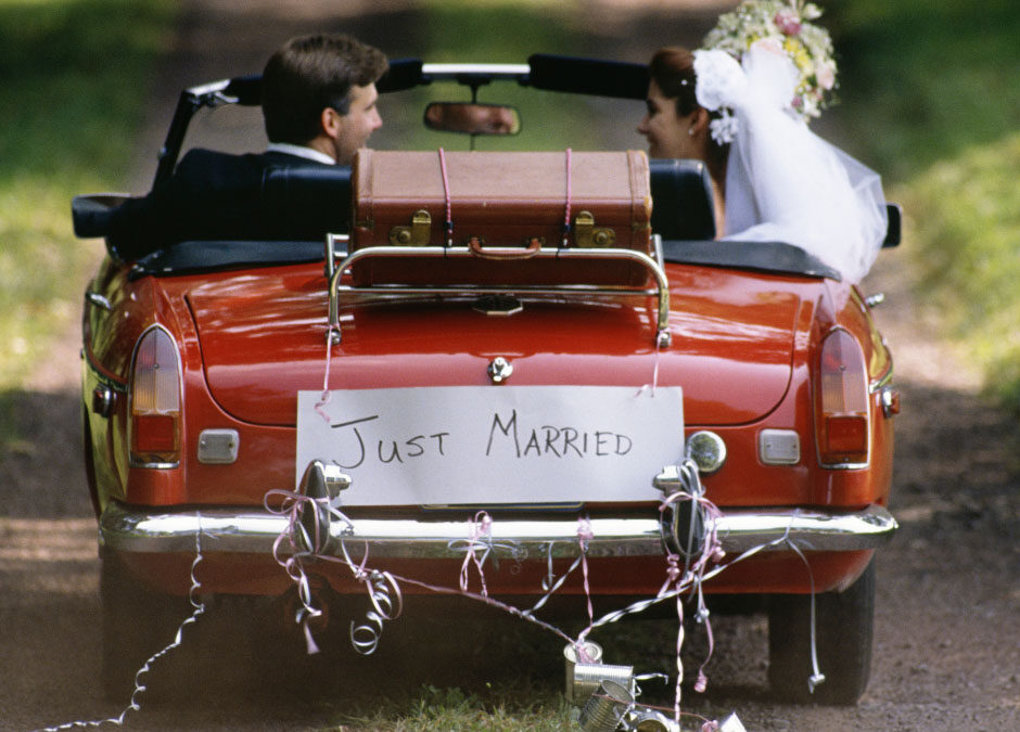 A newlywed couple smiles at each other while driving away in a red convertible down a dirt road. A "Just Married" sign, ribbons, and trailing tin cans are attached to the rear bumper below a luggage rack holding a vintage suitcase. - mwkellyinsurance.com